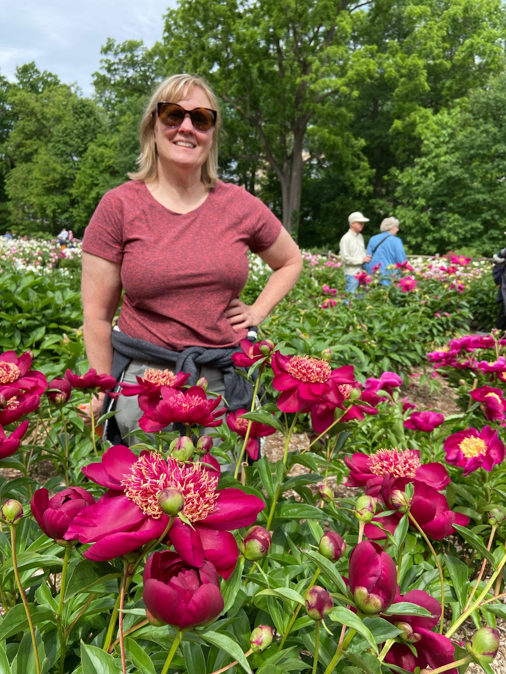 blonde woman named Laurie stands in garden of red peonies