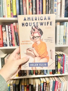 woman's hand holding American Housewife book in front of bookshelves